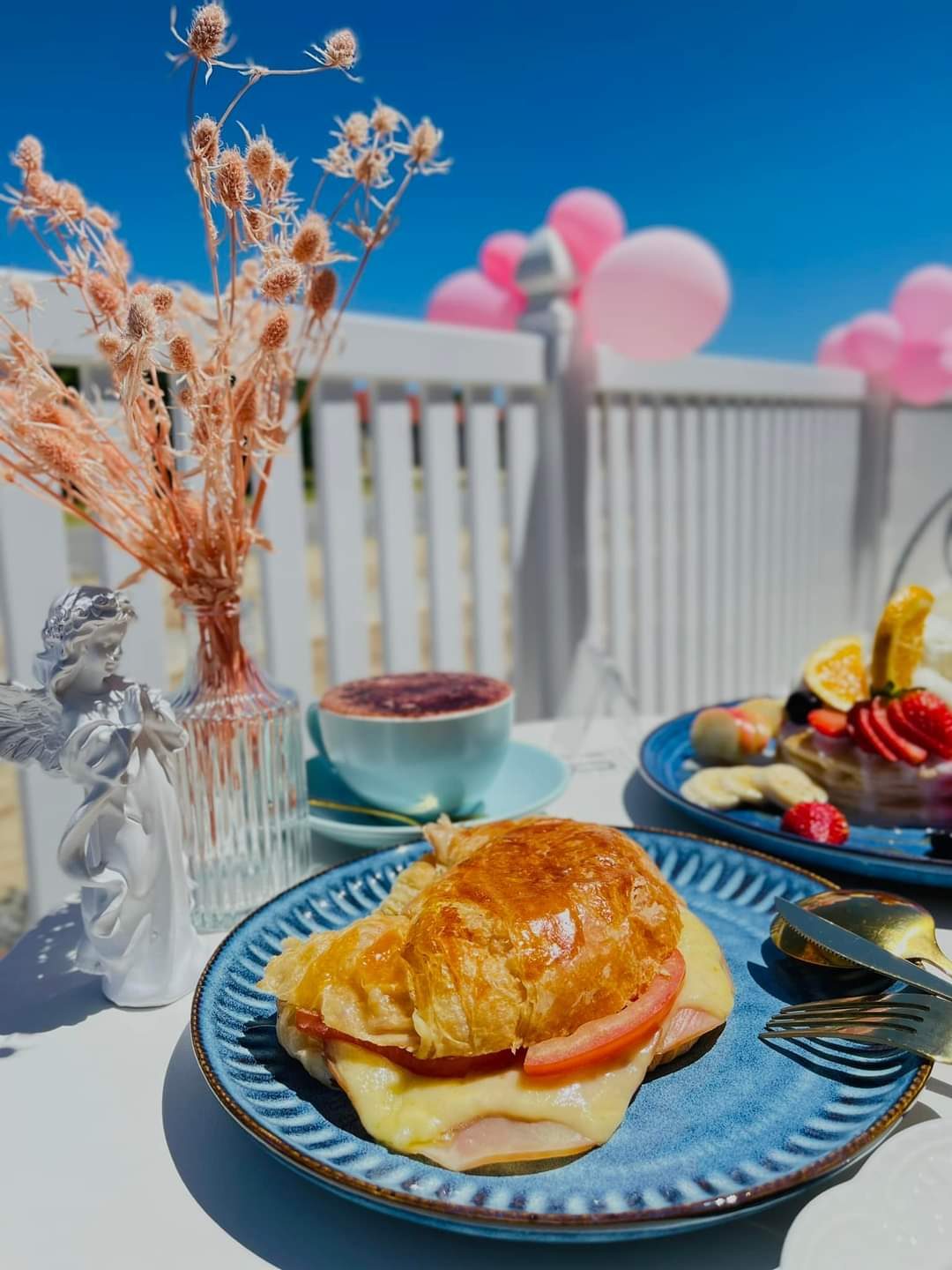 A ham, cheese and tomato croissant with a coffee on the outdoor table, pink balloons behind